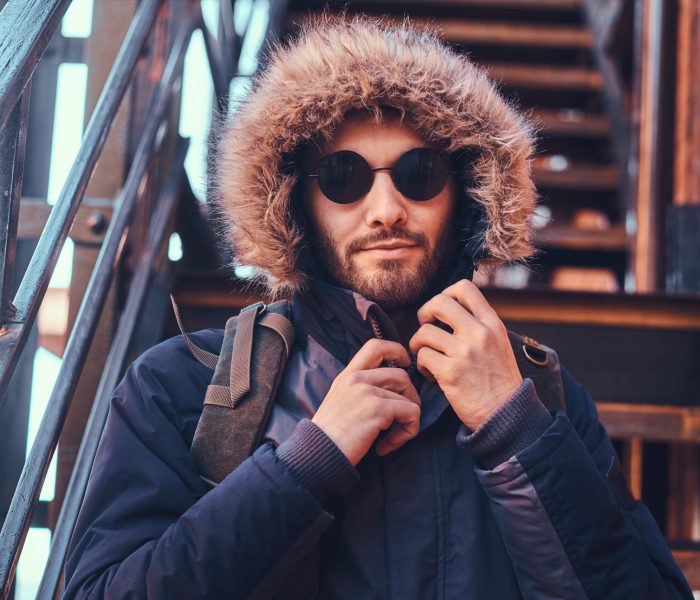 A handsome stylish young man wearing a winter coat and sunglasses sitting on stairs outside.